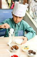 mignonne Indien chef garçon en train de préparer sundae plat comme une partie de non Feu cuisine lequel comprend vanille la glace crème, lutin, coco poudre, fraîchement haché des fruits et fraise sirop. peu enfant en train de préparer nourriture photo