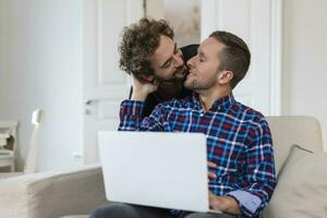 charmant gay couple en riant ensemble tandis que séance dans leur vivant pièce à maison. deux romantique Jeune Masculin les amoureux ayant amusement surfant le l'Internet à l'intérieur. Jeune gay couple vivant ensemble. photo