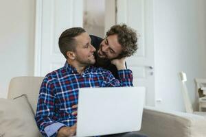 charmant gay couple en riant ensemble tandis que séance dans leur vivant pièce à maison. deux romantique Jeune Masculin les amoureux ayant amusement surfant le l'Internet à l'intérieur. Jeune gay couple vivant ensemble. photo