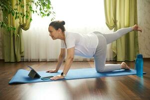 Jeune Enceinte femme en train de regarder en ligne grossesse yoga cours sur numérique tablette, Faire prénatal souimanga, oiseau chien pose photo