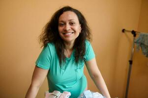 Enceinte femme, femme enceinte mère sourit à caméra, permanent par repassage planche avec repassé vêtements pour sa nouveau née bébé photo