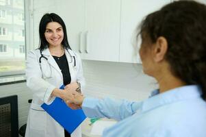 sur de soi caucasien femme médecin en portant presse-papiers avec médian Assurance contracter, souriant, tremblement main à Nouveau patient photo