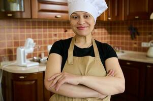 agréable femme au foyer dans du chef chapeau et tablier, permanent avec bras franchi dans le Accueil cuisine, souriant, à la recherche à caméra photo