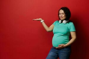 content souriant Enceinte femme dans décontractée vêtements et écouteurs, en portant copie un d espace sur sa main, isolé rouge toile de fond photo