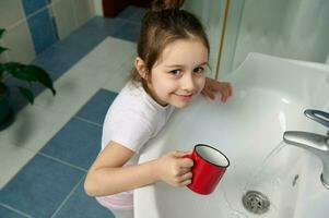 malicieux bébé fille permanent par évier dans Accueil salle de bains, souriant à la recherche à caméra tandis que nettoyage bouche, brossage les dents photo