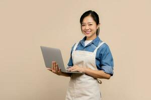 souriant Jeune asiatique femme barista barman employé portant marron tablier travail dans café magasin en utilisant travail sur portable PC ordinateur sur lumière marron Contexte. petit affaires Commencez concept photo