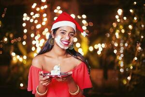 jolie content Jeune Indien femme dans Nouveau année chapeau en portant gâteau, crème brisé plus de sa visage photo