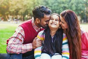 mignonne Indien peu fille étant embrassé par tous les deux de sa Parents sur le des joues Extérieur dans le parc photo