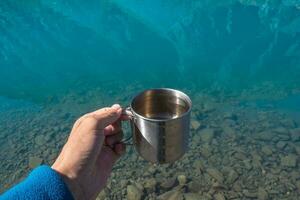 agresser avec pur glacial l'eau de une Montagne lac. main en portant une randonneurs tasse avec nettoyer en buvant l'eau. nettoyer en buvant l'eau dans le bleu Montagne lac, proche en haut. photo