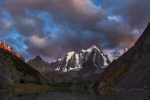 obscurité Montagne paysage avec génial neigeux Montagne allumé par Aube Soleil parmi foncé des nuages. impressionnant alpin paysage avec haute Montagne sommet à le coucher du soleil ou à lever du soleil. gros glacier sur Haut dans Orange lumière. photo