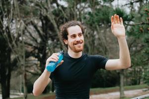 Un jeune homme athlétique sourit et salue tout en tenant une boisson gazeuse dans le parc tout en faisant du fitness