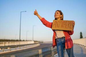 femme est auto-stop sur bord de la route en essayant à Arrêtez auto. elle est en portant papier carton avec une inscription. photo