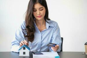 asiatique femme avec longue cheveux dans le bleu chemise est en utilisant une crédit carte pour achat une Nouveau maison et à la recherche à achat d'une maison les documents le idée de achat une Accueil avec une crédit carte. photo