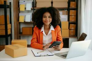 Commencez petit affaires PME, entrepreneur propriétaire africain femme en utilisant téléphone intelligent ou tablette prise recevoir et vérification en ligne achat achats commande à en train de préparer pack produit boîte. photo