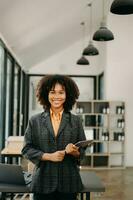 sur de soi africain femme avec une sourire permanent en portant bloc-notes et tablette à le bureau. photo