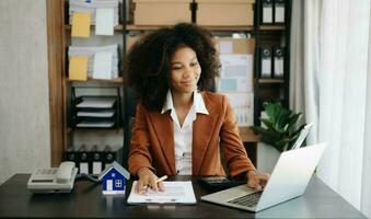 Jeune réel biens agent ouvrier travail avec portable et tablette à table dans Bureau et petit maison à côté de il. photo