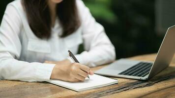 asiatique Jeune femme dans intelligent décontractée porter l'écriture sur carnet et travail sur portable tandis que séance dans Créatif Bureau ou café. Jeune fille travail avec portable sur le bois table dans le café photo