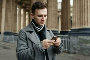 souriant Jeune homme dans gris manteau est en portant faire défiler texte messages dans le sien mobile photo