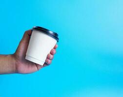 fermer Jeune adulte homme main en portant blanc tasse papier de café chaud boisson prêt à boisson rafraîchissant. arôme éveillé Frais à travail mis sur une bleu isolé Contexte. point de vue tir. photo