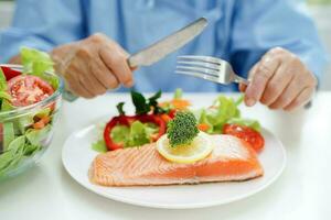 asiatique personnes âgées femme patient en mangeant Saumon pieu et légume salade pour en bonne santé nourriture dans hôpital. photo