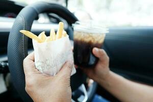 asiatique femme chauffeur tenir et manger français frites dans voiture, dangereux et risque un accident. photo