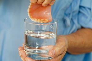 asiatique Sénior femme patient nettoyer les dents dentier dans une verre avec Solution pour mâcher aliments. photo