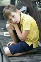 triste peu garçon est assis sur une parc banc. photo