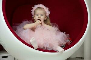deux ans enfant. une intelligent peu fille dans une rose duveteux robe est assis sur une rouge chaise. photo