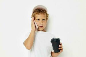 peu garçon dans une blanc T-shirt casquette avec une téléphone dans une verre avec une boisson lumière Contexte inchangé photo