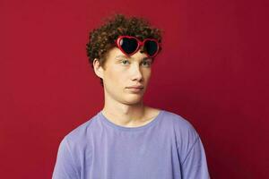 portrait de une Jeune frisé homme gars dans une violet T-shirt avec des lunettes de soleil posant isolé Contexte photo