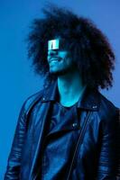 portrait de mode homme avec frisé cheveux avec élégant des lunettes sur bleu Contexte multinational, coloré lumière, noir cuir veste s'orienter, moderne concept. photo