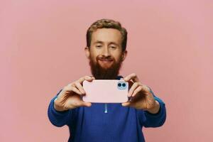 portrait de une rousse homme avec téléphone dans main prise selfies et Photos sur le sien téléphone avec une sourire sur une rose arrière-plan, blogueur