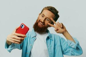 homme branché avec téléphone dans main blogueur sourire sur gris Contexte dans bleu chemise et blanc T-shirt, parlant sur le téléphone et selfies photo