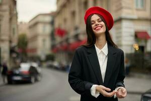 mode femme portrait sourire les dents permanent sur le rue dans le ville Contexte dans élégant vêtements avec rouge lèvres et rouge béret, voyage, cinématique couleur, rétro ancien style, Urbain mode mode de vie. photo