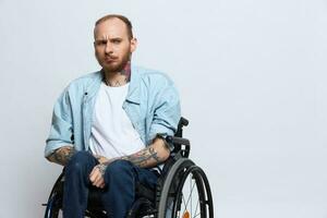 une homme dans une fauteuil roulant regards à le caméra, avec tatouages sur le sien bras est assis sur une gris studio arrière-plan, le concept de santé est une la personne avec handicapées photo