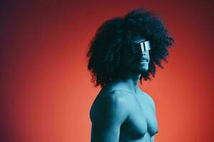 portrait de mode homme avec frisé cheveux sur rouge Contexte avec élégant lunettes, multinational, coloré lumière, noir cuir veste s'orienter, moderne concept, sexy corps. photo