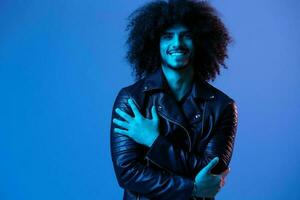 portrait de mode homme avec frisé cheveux sur bleu Contexte multinational, coloré lumière, noir cuir veste s'orienter, moderne concept. photo