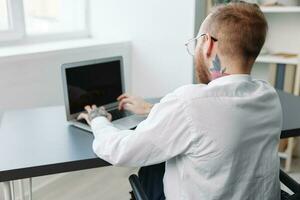 une homme fauteuil roulant homme d'affaire avec tatouages dans le Bureau travaux à une portable en ligne, affaires processus, une fauteuil roulant fermer, l'intégration dans société, le concept de travail une la personne avec handicapées photo