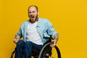 homme dans une fauteuil roulant cris avec colère, spectacles poing regards à le caméra est ne pas heureux, avec tatouages sur le sien mains est assis sur Jaune Contexte de le studio, le concept de santé est la personne avec handicapées photo