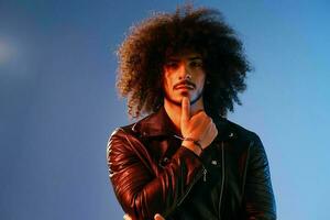 portrait de une élégant homme avec frisé cheveux sur une bleu Contexte multinational, coloré lumière, noir cuir veste s'orienter, moderne concept. photo