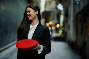 mode femme sourire avec les dents et amusement touristique dans élégant vêtements dans veste et rouge béret en marchant vers le bas étroit ville rue en volant cheveux, voyage, français style, cinématique couleur, rétro ancien style. photo