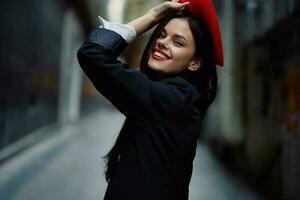 mode femme sourire avec les dents et amusement touristique dans élégant vêtements dans veste et rouge béret en marchant vers le bas étroit ville rue en volant cheveux, voyage, français style, cinématique couleur, rétro ancien style. photo