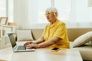 content personnes âgées femme avec une portable dactylographie dans écouteurs sourire séance à Accueil sur le canapé dans une Jaune chemise, brillant moderne intérieur, mode de vie en ligne communication. photo