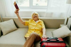 content Sénior femme avec passeport et Voyage billet emballé une rouge valise, vacances et santé se soucier. souriant vieux femme joyeusement séance sur le canapé avant le voyage élevé sa mains en haut dans joie. photo