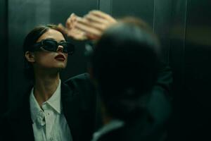 élégant femme dans une noir veste et des lunettes de soleil posant dans un ascenseur ascenseur près le miroir, mode modèle, foncé cinématique lumière et Couleur matrice, charme ancien photo