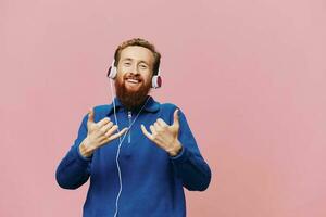portrait de une rousse homme portant écouteurs souriant et dansant, écoute à la musique sur une rose Contexte. une branché avec une barbe. photo