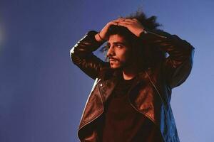 portrait de une élégant homme avec frisé cheveux sur une bleu Contexte multinational, coloré lumière, noir cuir veste s'orienter, moderne concept. photo