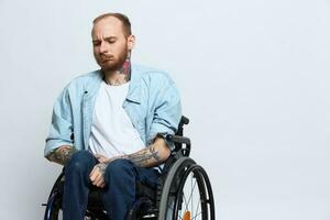 une homme dans une fauteuil roulant regards à le caméra, avec tatouages sur le sien bras est assis sur une gris studio arrière-plan, le concept de santé est une la personne avec handicapées photo