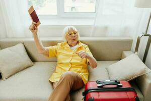 content Sénior femme avec passeport et Voyage billet emballé une rouge valise, vacances et santé se soucier. souriant vieux femme joyeusement séance sur le canapé avant le voyage élevé sa mains en haut dans joie. photo