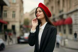 mode femme portrait sourire avec les dents permanent sur le rue dans le ville Contexte dans élégant vêtements rouge lèvres et rouge béret, voyage, cinématique couleur, rétro ancien style, Urbain mode mode de vie. photo